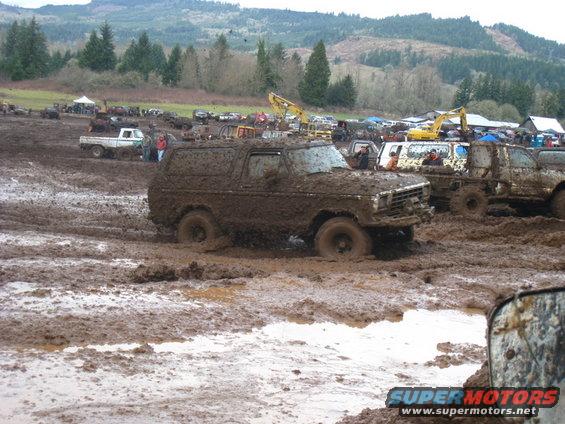 1978 Ford Bronco stuck deep in mud, other vehicles and trees in background