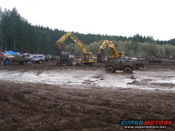 Muddy field with two excavators, a monster truck, and several off-road vehicles. People are gathered around some trucks