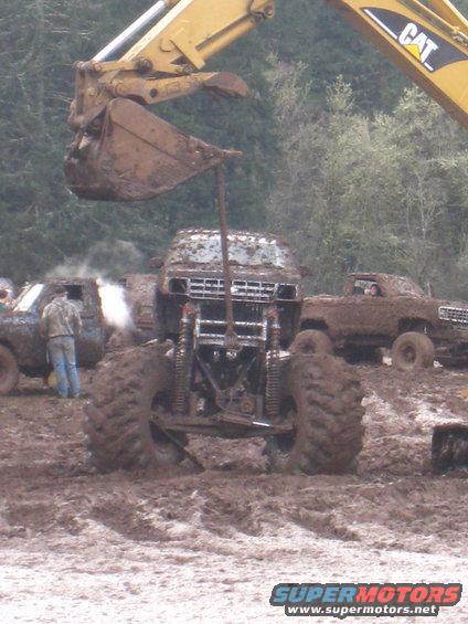 Monster trucks covered in mud, one being lifted by an excavator bucket