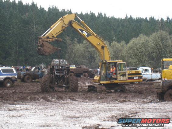 Excavator digging in mud pit with several modified trucks around