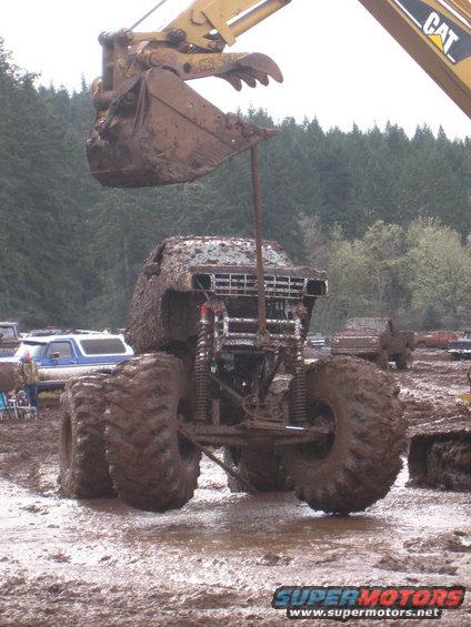 Monster truck stuck in mud, excavator bucket overhead. Other trucks in background