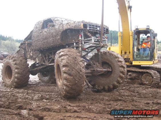 Mud-covered 1978 Ford Bronco monster truck with large tires, next to a yellow excavator