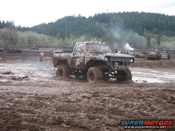 Mud-covered pickup truck stuck in mud pit, other vehicles in background