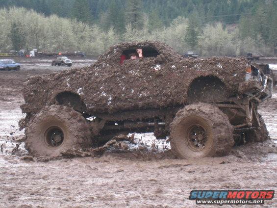 Mud-covered off-road vehicle with large tires stuck in mud, driver visible inside