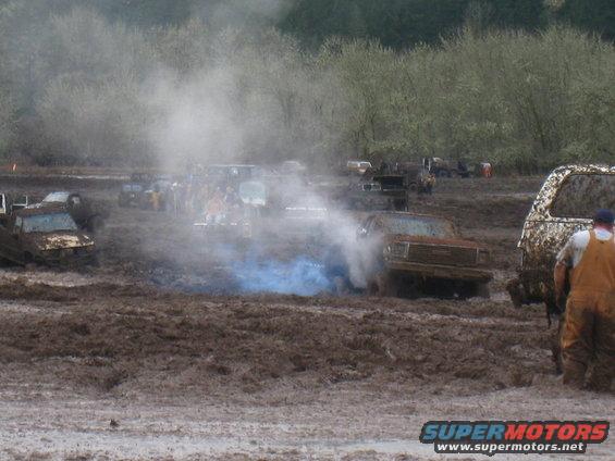 Mud bogging scene with trucks stuck, one emitting blue smoke