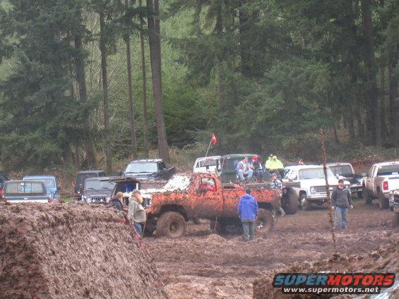 Muddy trucks and people gathered in a wooded area with a large pile of mud in the foreground