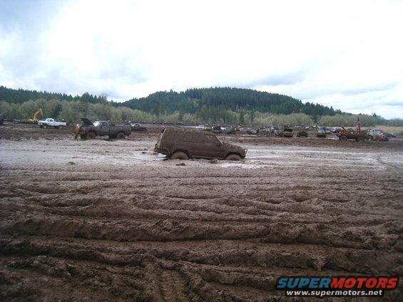 SUV stuck deep in mud pit, other trucks and vehicles visible in background