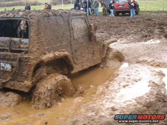 Mud-covered off-road vehicle stuck in mud pit, other vehicles and spectators in background