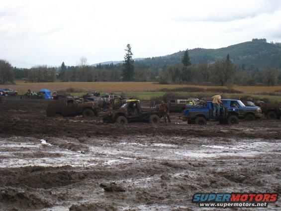 Several trucks and SUVs stuck in a muddy field with trees and hills in the background