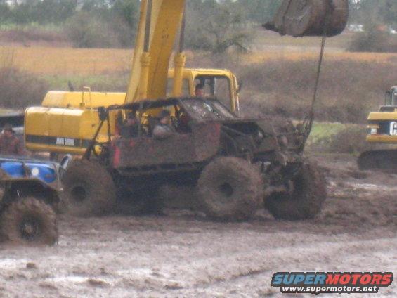 Mud-covered off-road vehicle with roll cage, large tires, and people inside, in a muddy field with excavators