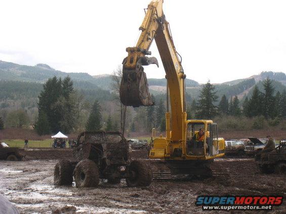 Excavator lifting a heavily mud-covered vehicle in a muddy field with trees and hills in the background
