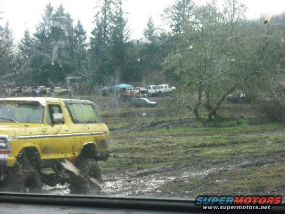 Yellow 1978 Ford Bronco with large tires splashing through mud. Other vehicles and trees in background