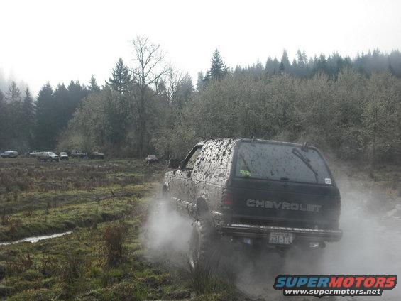 Muddy Chevrolet SUV kicking up dust on a dirt track with trees and other vehicles in the background
