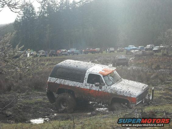 Mud-covered white and orange SUV stuck in mud, with other vehicles in background