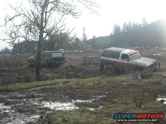 Two SUVs, one black and one white with orange stripes, stuck in a muddy field. A tow rope connects them