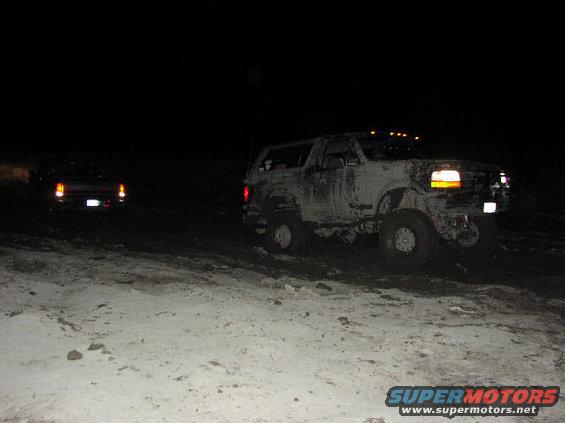 Mud-covered 1992 Ford Bronco with roof lights driving off-road at night
