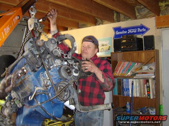 Man in plaid shirt working on a blue engine suspended by an orange engine hoist