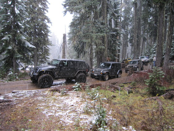 Black Jeep Wrangler with large tires on a muddy, snow-dusted forest road, 3/4 front view