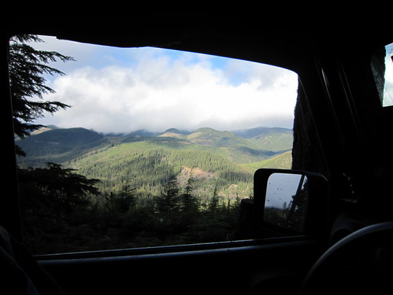 View from inside a dark vehicle looking out a window at a misty mountain range covered in pine trees. A side mirror is visible
