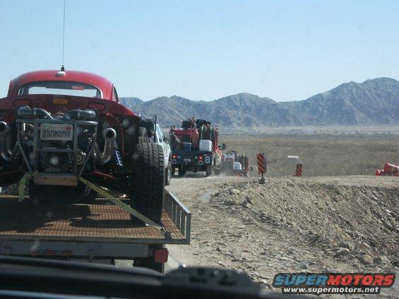 Red custom dune buggy with exposed engine on a trailer, driving in a convoy on a dirt road with mountains in the background