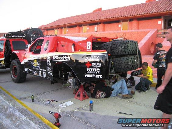 Off-road race truck with BFGoodrich tires and various sponsor decals, being worked on by mechanics. Another vehicle is parked behind it