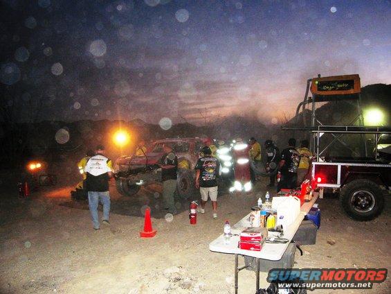 Red off-road vehicle with modified front end and large tires, surrounded by people and equipment at dusk