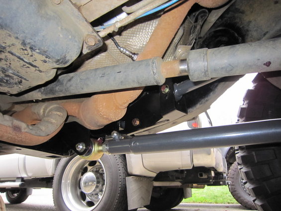 Undercarriage of a vehicle showing suspension components, exhaust, and driveshaft. Two other trucks are partially visible