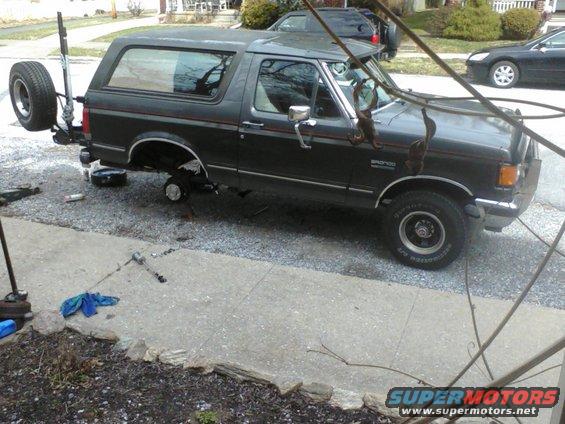 Dark gray Ford Bronco with spare tire mounted on the back, front wheel removed, and jack stands under the rear