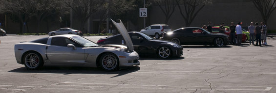 Silver Corvette with hood open, parked next to a black sports car and a white SUV. Group of people stand near other cars in background