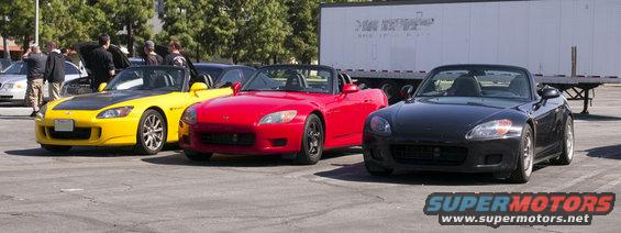 Three Honda S2000 convertibles parked in a lot, one yellow, one red, one black. A semi-trailer is behind them