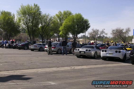 Silver and white sports cars parked in a lot, rear view. Some people stand around