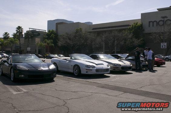Several sports cars, including a black Corvette and a white convertible Camaro, parked in a lot with buildings in the background