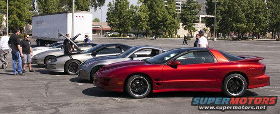 Red Pontiac Firebird and silver sports cars lined up in a parking lot. People are standing around them