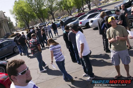 People gathered around cars in a parking lot. Some are looking at cars, others are talking