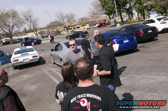 People gathered around cars in a parking lot, including a white classic muscle car and a blue sedan