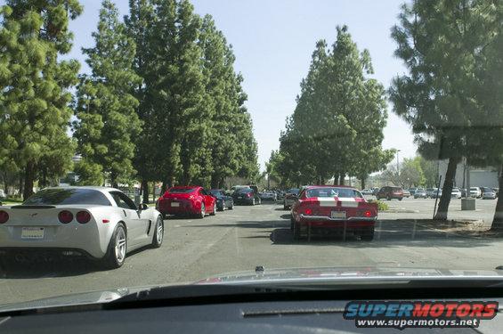 Several cars driving in a line, including a silver Corvette and a red classic muscle car