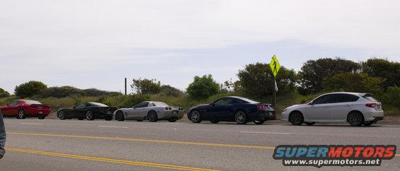 Several cars, including a red Dodge Challenger, two Chevrolet Corvettes, a Ford Mustang, and a Subaru Impreza, parked in a line along a roadside