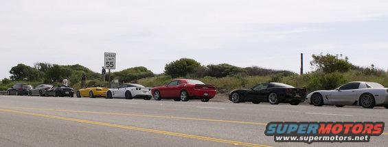 Several sports cars parked on the side of the road, including a red Dodge Challenger and multiple Chevrolet Corvettes. A speed limit sign is visible