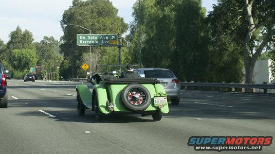 Green vintage convertible with spare tire on rear, driving on highway. Silver car behind it