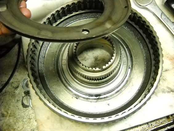 Close-up of a transmission component with gears and a ring being held above it. A wrench is visible in the background