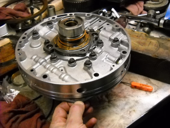 Automatic transmission pump assembly with bolts and internal components visible. A hand is holding the edge