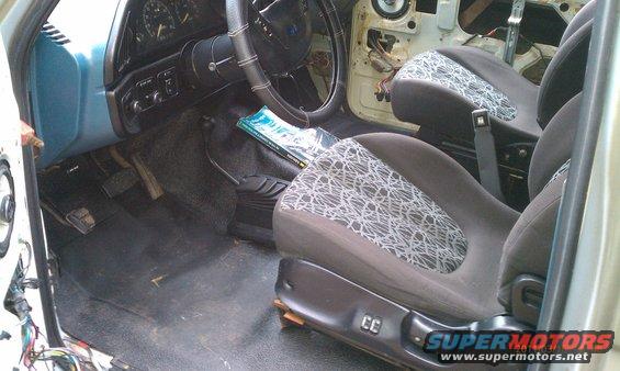 Interior of a vehicle, showing seats, dashboard, steering wheel, and floor pedals. Door panel is removed