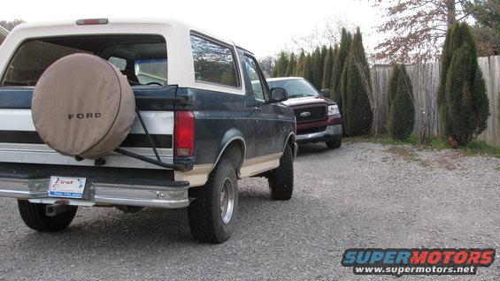 Rear view of a dark blue and tan Ford Bronco with a spare tire on the back, parked next to a red Ford F-150