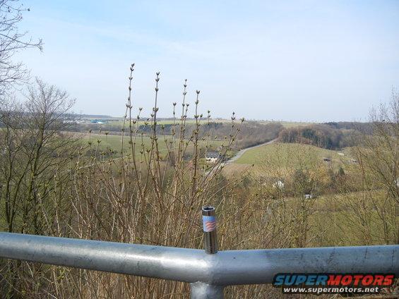 Metal railing in foreground with a lighter on it, overlooking a rural landscape with fields and trees under a blue sky