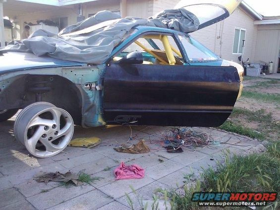 Car with door open, missing fender, and exposed wiring, parked on a paved surface