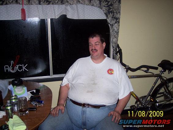 Man in white t-shirt and jeans sits at a table with drinks; bicycle behind him. Neon sign in window reads 'ROCK'