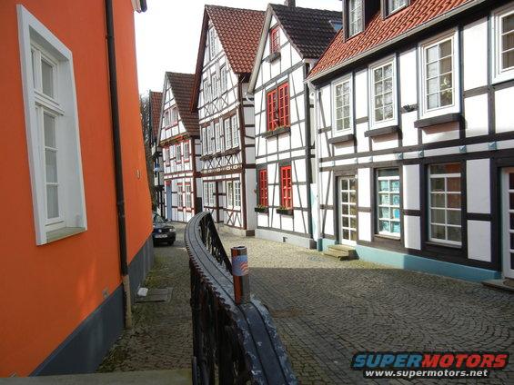 Narrow cobblestone street lined with traditional German half-timbered houses, an orange building on the left, and a dark car parked on the right