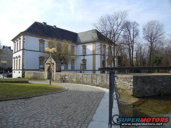 Cobblestone path leads to a large white building with a dark roof, surrounded by trees and a stone wall