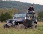 Dark green Jeep Wrangler with a person on top, parked in a grassy field