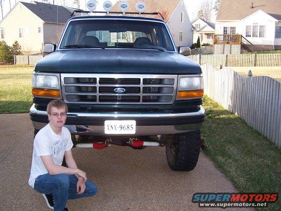 Guy crouching in front of a dark green Ford Bronco. It has a roof rack with lights and a license plate reading XNX-9685